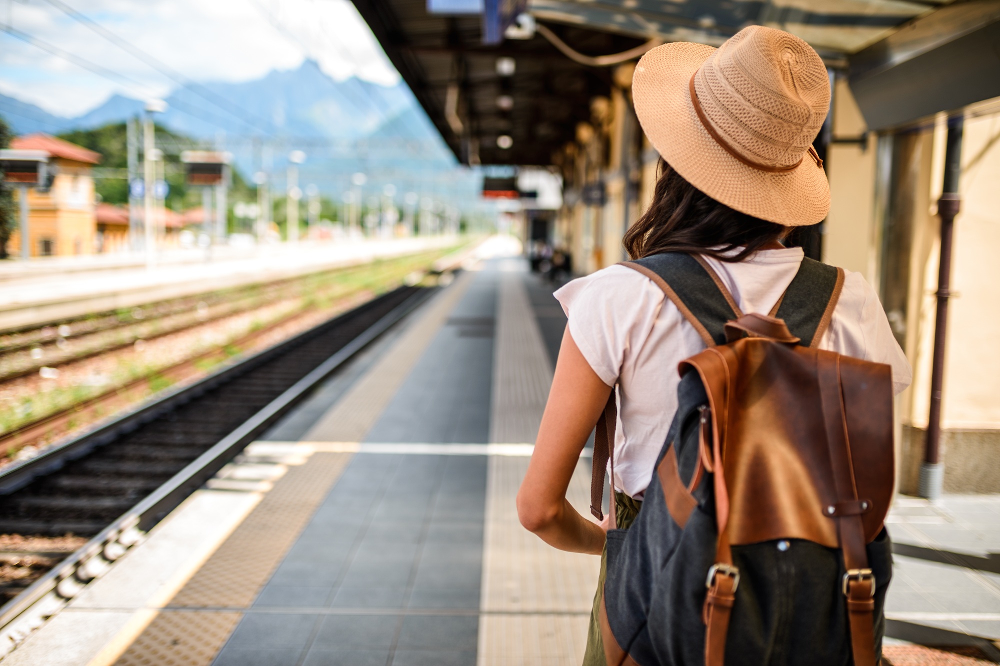 Eine sommerlich gekleidete Frau mit einem Strohhut steht am Bahngleis und wartet auf einen Zug